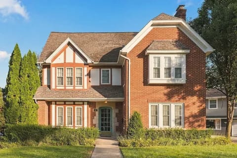 A two-story brick Tudor-style house with a front walk between trimmed hedges.